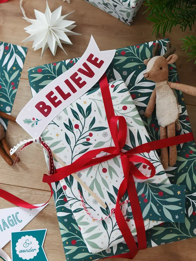 A stack of Christmas gifts wrapped in botanical wrapping paper with a red and white striped ribbon lay on a wooden floor. A Believe flag is tucked into the ribbon, and a small reindeer toy lays beside it.