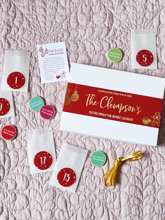 A white advent calendar box with a red sleeve lays on a pink quilt with white numbered envelopes beside it.