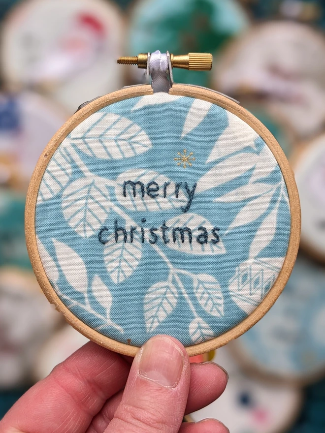 A hand embroidered hoop on Christmas fabric reading merry christmas