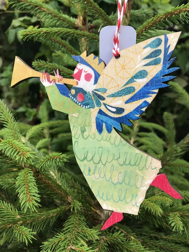 Joyful Angel with trumpet wooden illustrated decoration by Esther Kent, hangs against the fresh green of a fir tree Christmas tree.