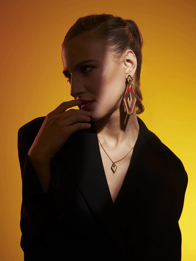 A woman modelling a hexagon shaped earrings in warm tones. The background is warm orange colour. The model is wearing black suit and her hand on the mouth and her hair is on a pony tail. 