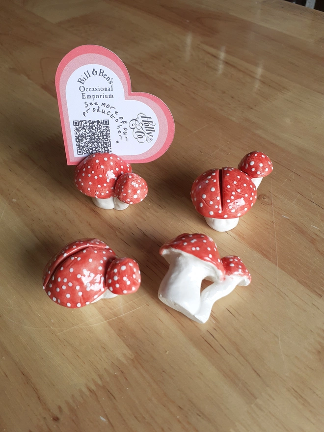 Set of four handmade ceramic name card holders shaped like clusters of red toadstools with white spots, displayed on a wooden table, with one holding a heart-shaped card.