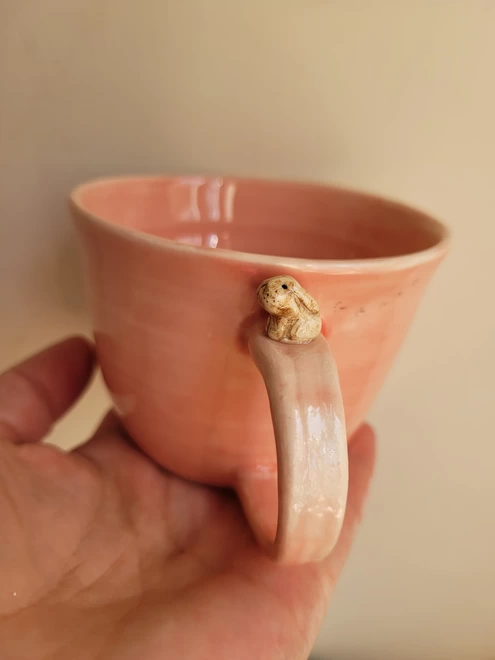 pink pottery cup is held in a hand it has a smll modelled brown bunny rabbit sitting on the handle and small footprints behind