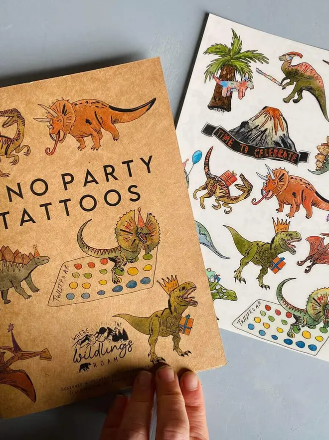 Dinosaur & Fantasy Temporary Tattoos, a hand holds a brown kraft paper booklet titled "DINOSAUR TATTOOS" next to a tattoo sheet with colorful dinosaur illustrations.
