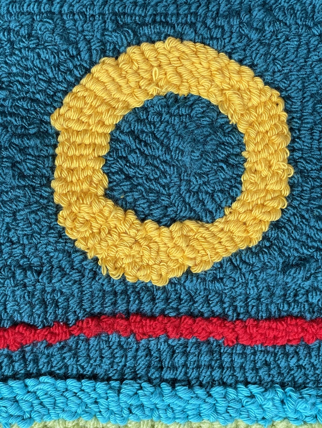 Purple wool loops making an "O" on a blue loop background with pink stripe underneath