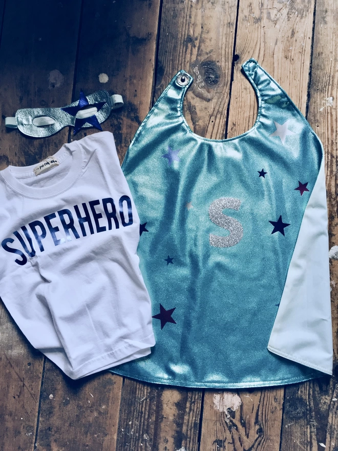 flatlay of personalised ice blue superhero costume - ice blue personalised cape, blue superhero t-shirt, ice & blue sapphire metallic leather Star superhero mask 