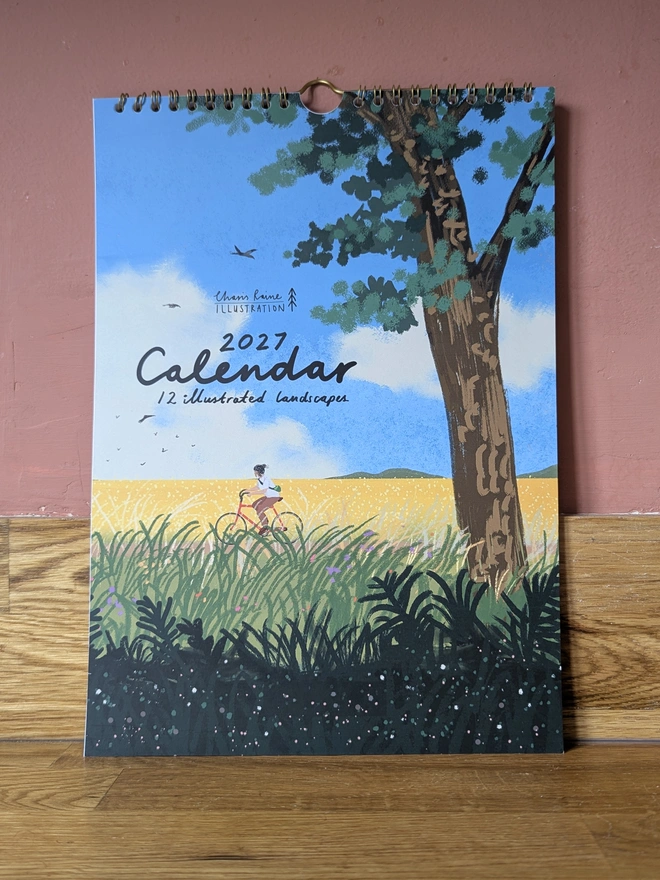 Illustrated landscape calendar by Charis Raine featuring a cyclist though yellow and green fields, with a tree and blue sky

