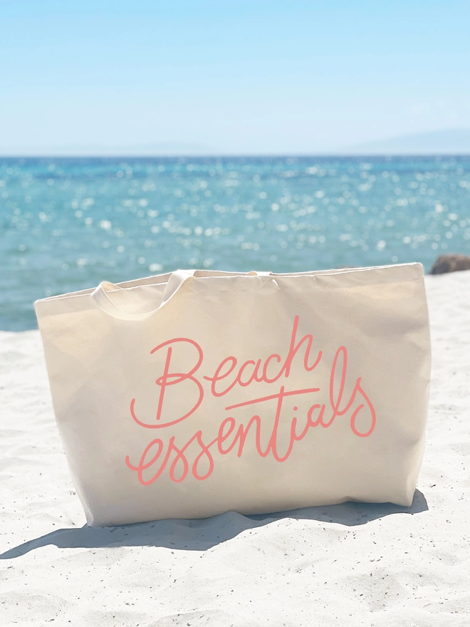 oversized natural canvas tote bag with beach essentials slogan on a beach with the sea in the distance