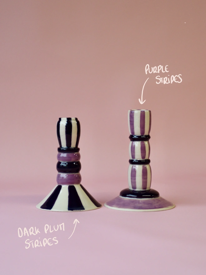 Handmade ceramic candlesticks with purple stripe patterns, including a dark plum striped design on the left and a lighter purple striped candlestick on the right, set against a pink background.