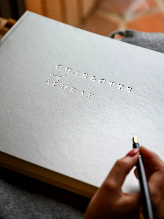 Landscape Wedding Guest Book, covered in Ivory Jewel Bookcloth, with a pen held ready to write inside.