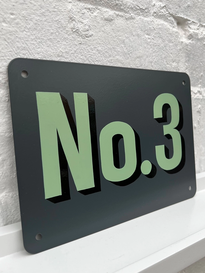 Sage green and black house number No. 3, on anthracite grey metal plaque, against a white brick wall.