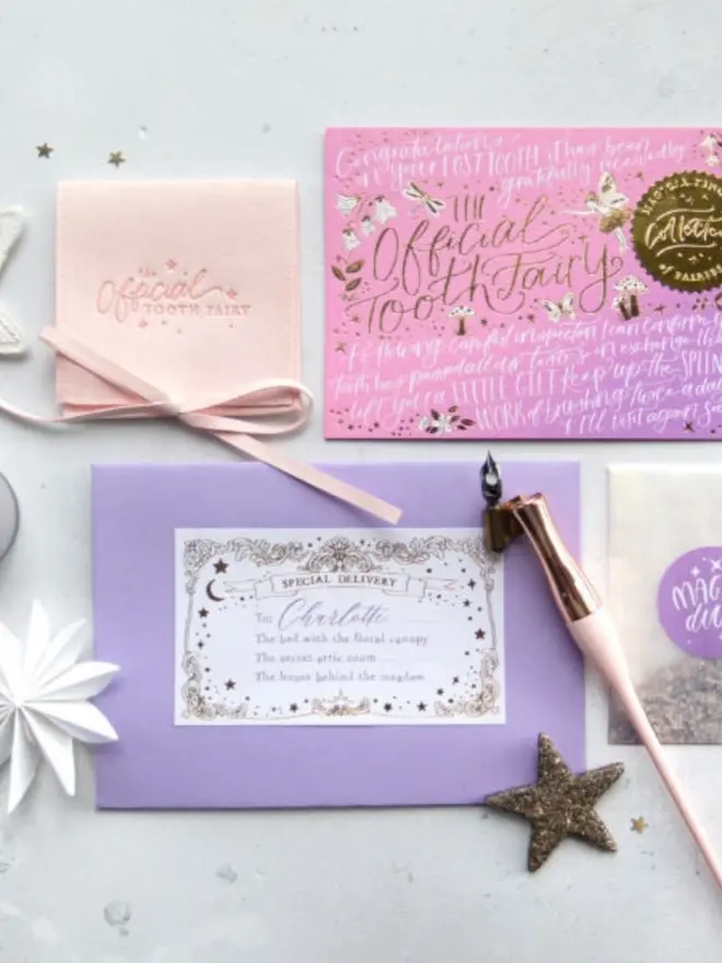 The pink-lilac notecard and a blush pink pouch sit above a lilac envelope addressed to Charlotte. A rose gold dip pen and a Magic Dust packet lie alongside.