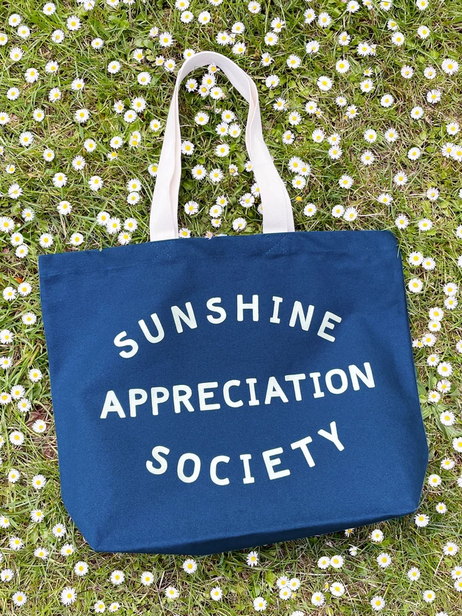 a large blue canvas tote bag with the words sunshine appreciation society on laying on the grass