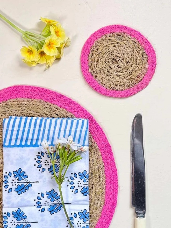 Seagrass coaster with pink rings, shown on a light surface with yellow  flowers. 