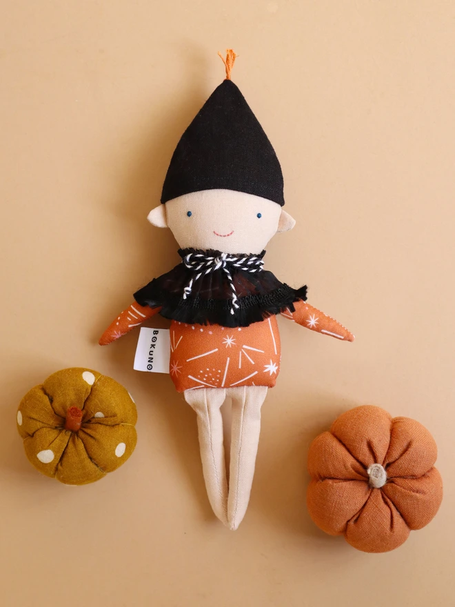 Handmade autumn elf doll with a black pointed hat, orange patterned top, black ruffled collar with black-and-white twine tie, embroidered blue eyes and pink smile, and light coloured face and legs 