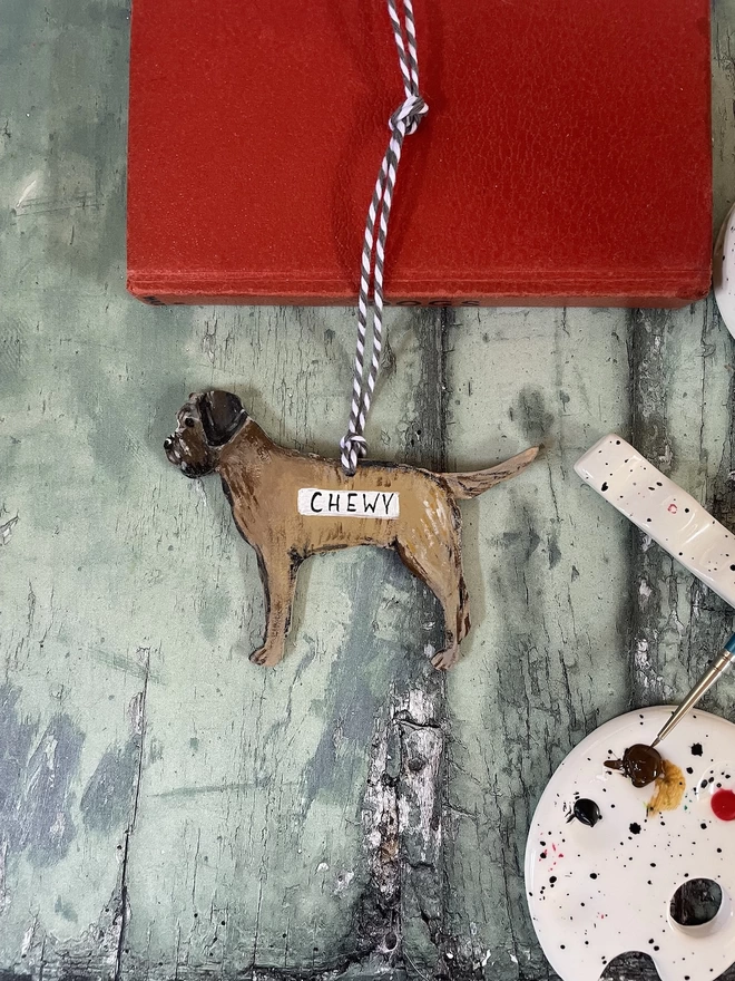 A hanging Border Terrier Decoration placed next to a paint palette with a paint brush resting nearby 