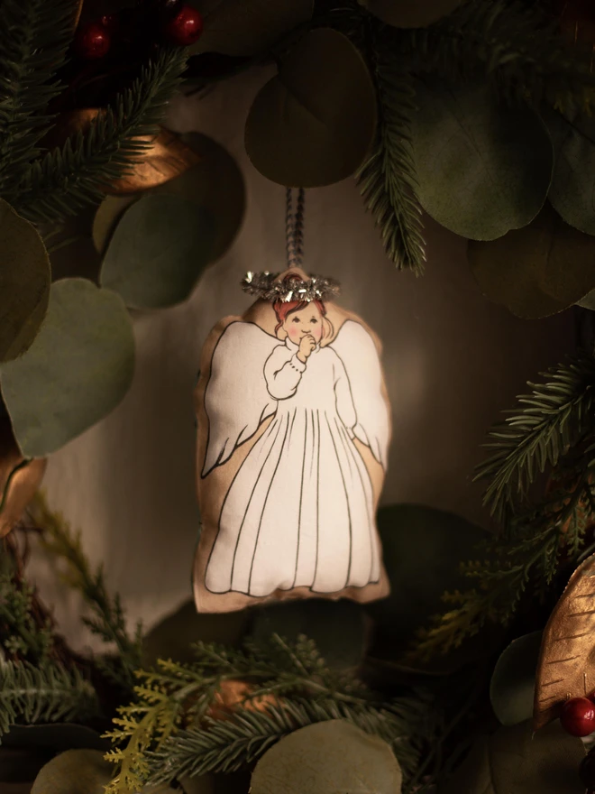 A Christmas tree angel decoration depicting a little girl angel sucking her thumb with red hair hung in the middle of a Christmas wreath