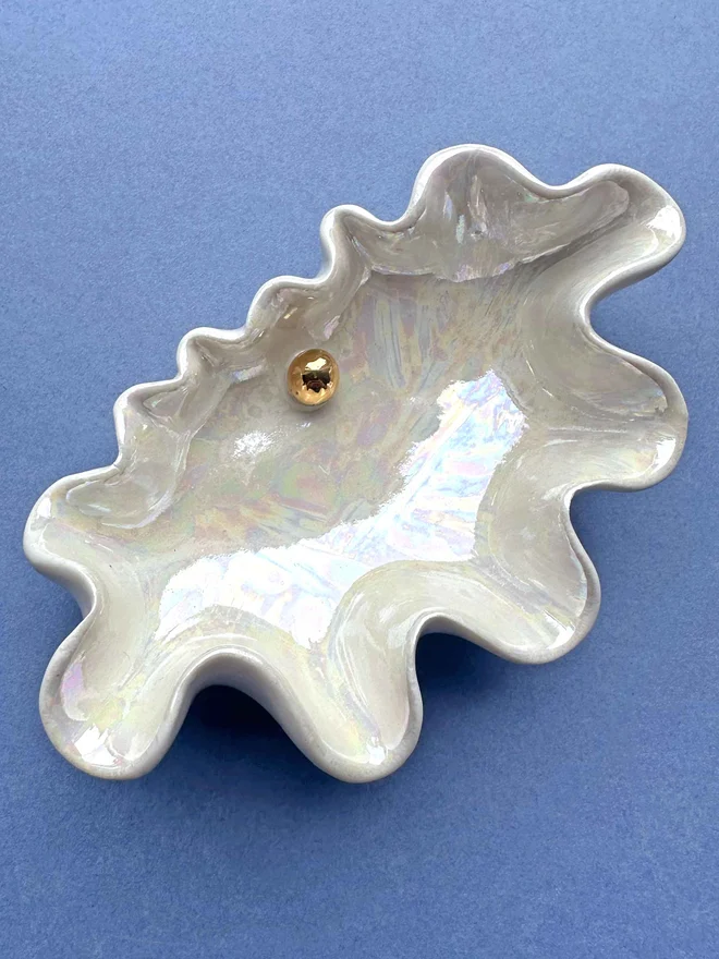 Porcelain clam shell pinch pot in shimmery white with gold mother of pearl in the middle of the shell, on a blue background