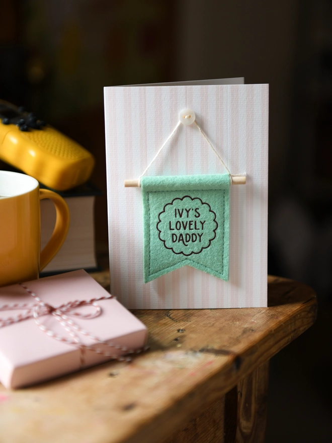 Handmade Father’s Day card with personalised green felt banner “Ivy's Lovely Daddy”, miniature hanging flag keepsake on wooden table. The flag is hung on the card from a tiny mother of pearl button. Beside the card is a book, a mug, a wrapped gift.
