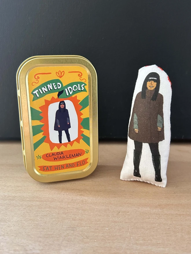 Mini fabric doll featuring an illustration of Claudia Winkleman in her Traitors cape. The doll is beside a gift tin.