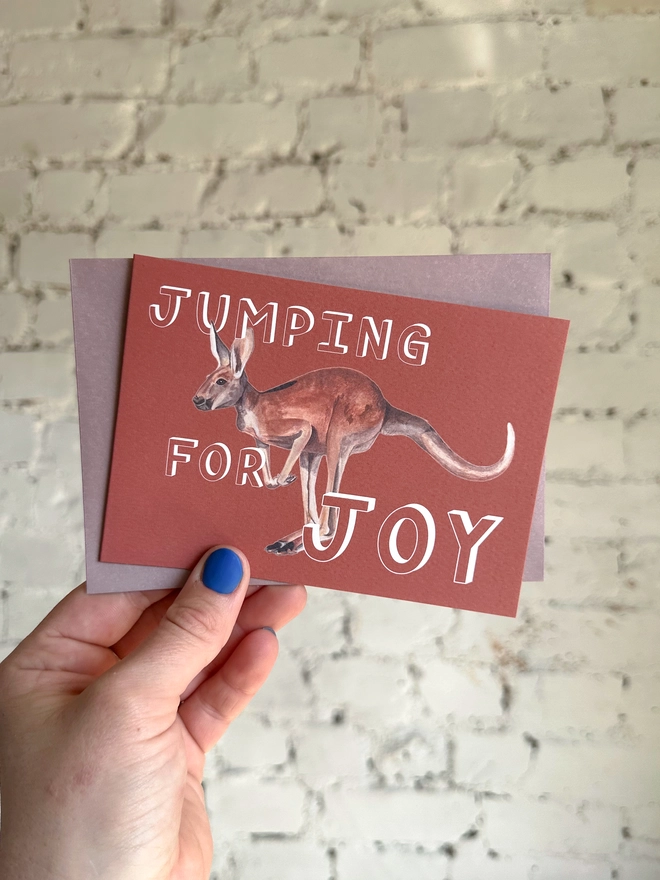 a rusty red greetings card featuring an illustration of a kangaroo with the text “jumping for joy” with a muted purple envelope