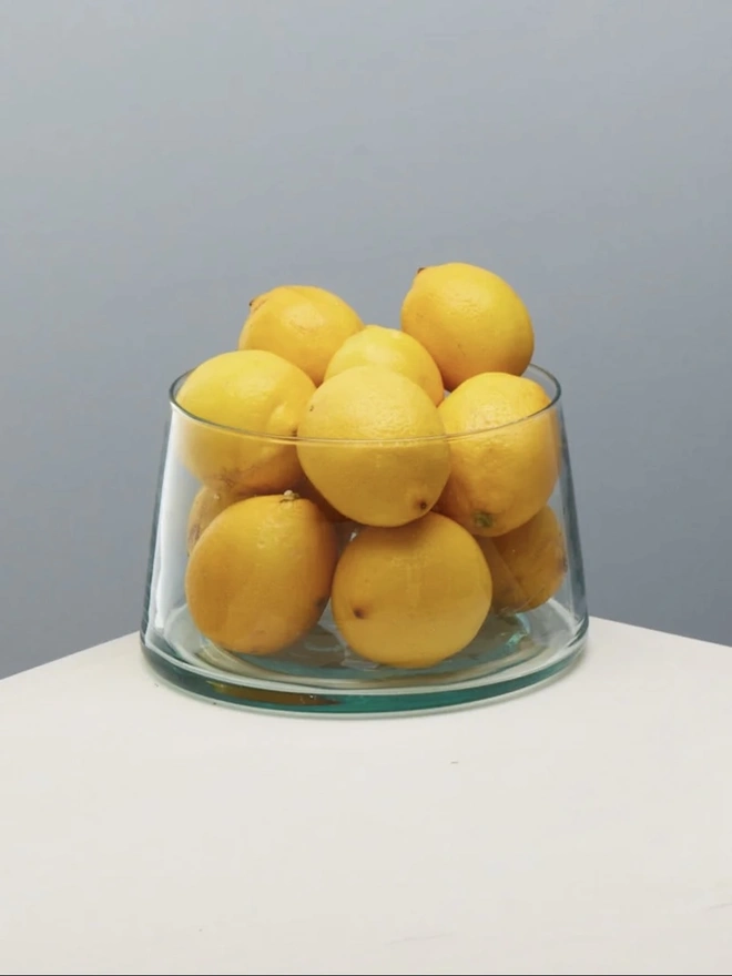 Tall Glass Dish, a tall glass dish sitting on a white surface against a baby blue backdrop. It is filled by lemons. 