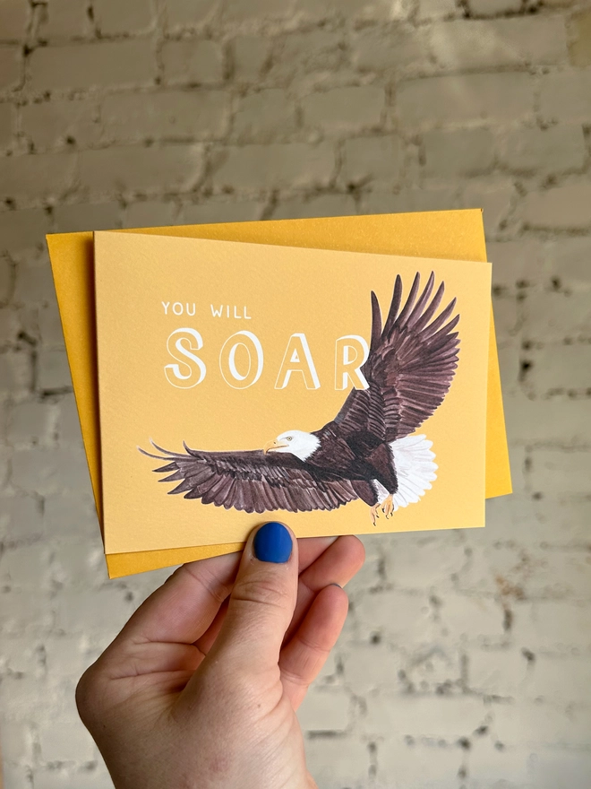 a bright yellow greetings card featuring an illustration of a bald eagle with the text “you will soar” with a deep yellow envelope