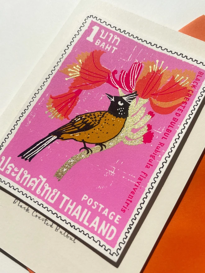 Bird Stamp Card Thailand Black Crested Bulbul, a white card with a colourful bird design in the centre on top of an orange envelope on a white surface. 