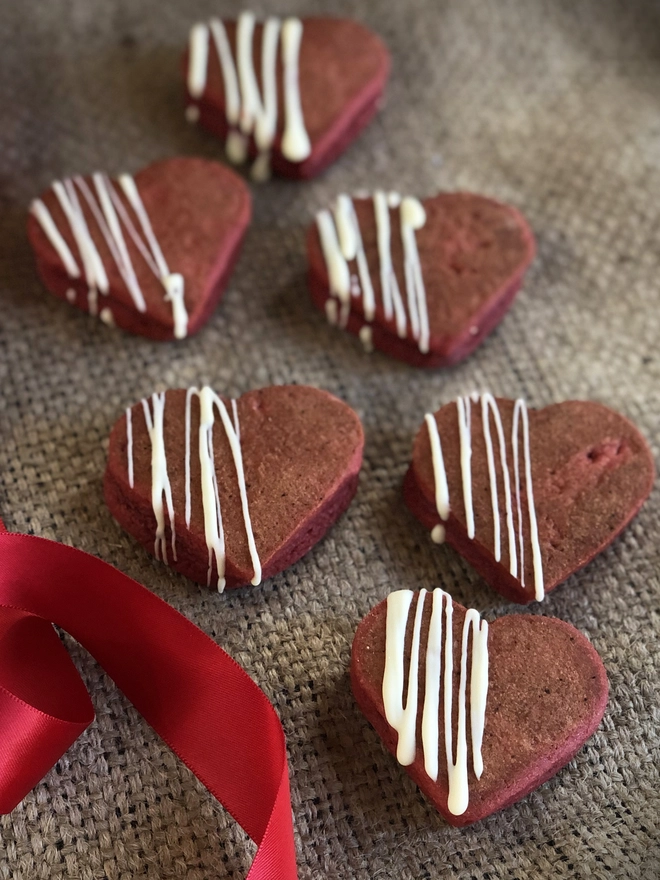 White chocolate topped, Red velvet heart shaped Welshcakes on a hessian top with a red ribbon laid on the left side.