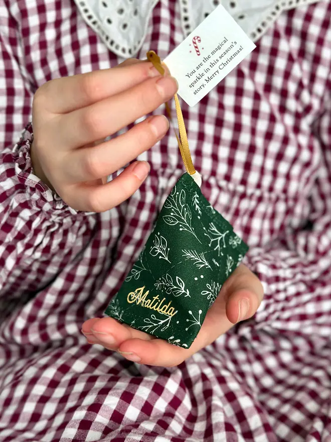 A child wearing a red gingham dress holds a green greenery personalised fabric fortune cookie with the name “Matilda” in gold, revealing a tiny wish card inside that reads “You are the magical sparkle in this season's story… Merry Christmas!”