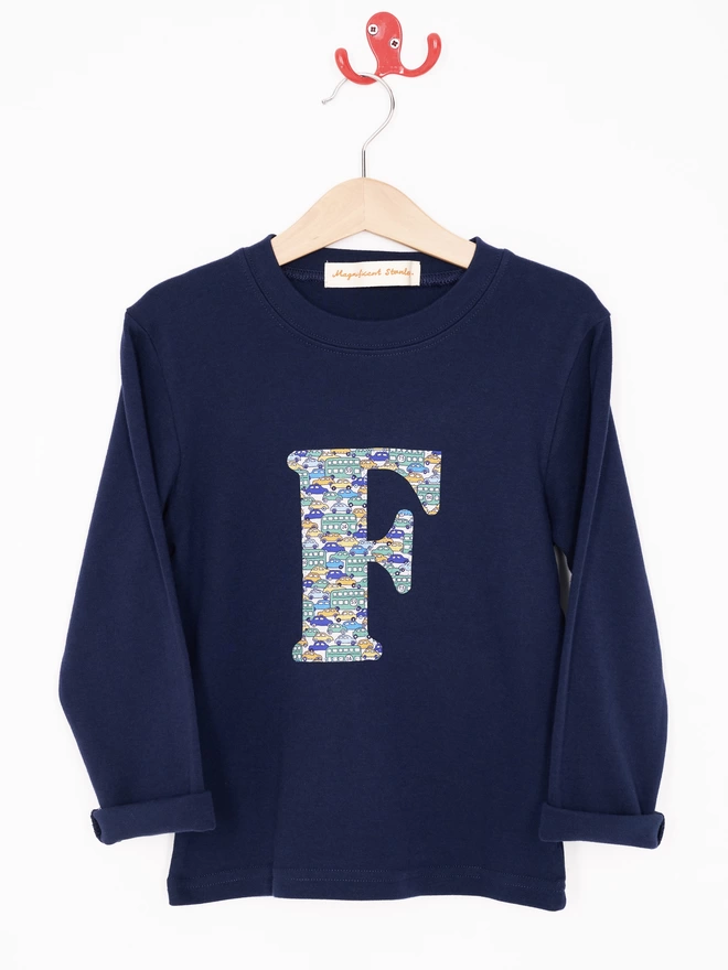 A navy cotton long sleeve t-shirt appliquéd with an initial in a vintage cars and busses Liberty print, hanging on a hanger