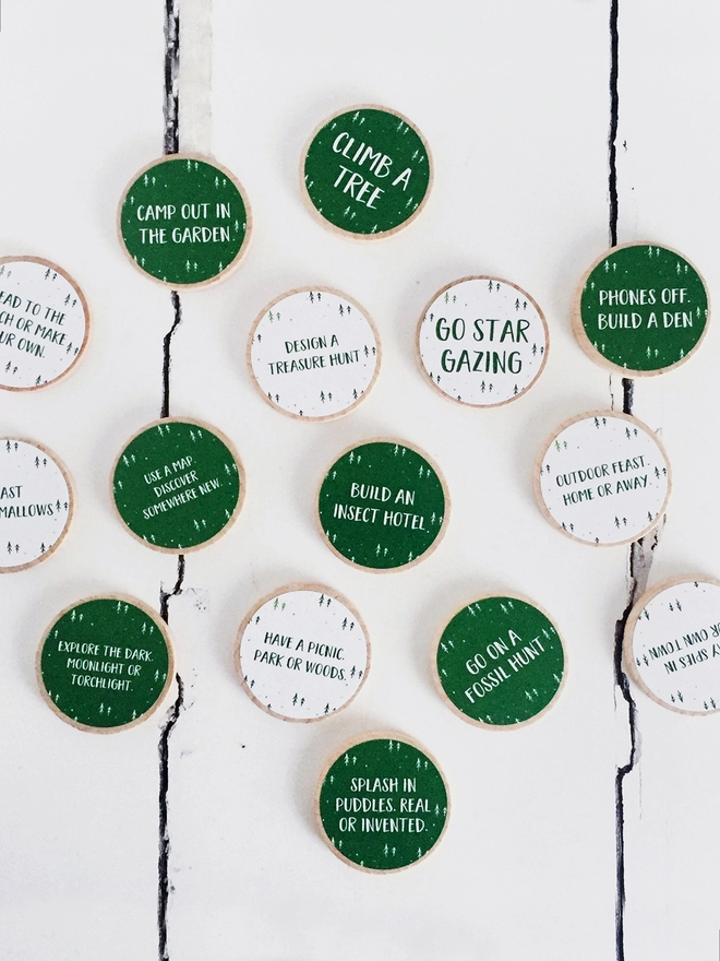 Fifteen wooden tokens with white and green labels lay on a white wooden surface. Each token has an outdoor adventure idea printed on.