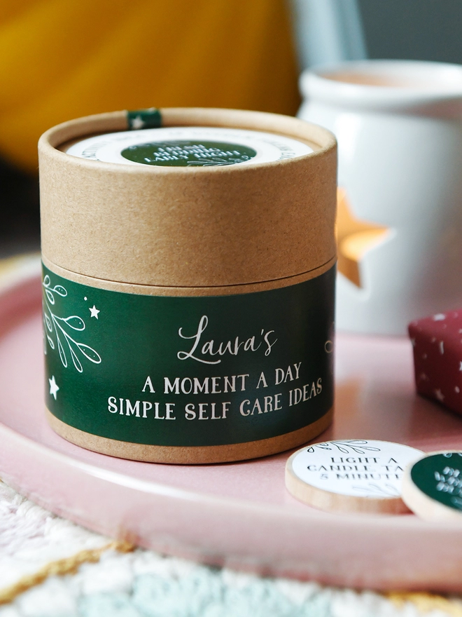 A cardboard jar with a dark green label that reads A moment a day, Simple self care ideas, stands on a pink plate beside wooden tokens.