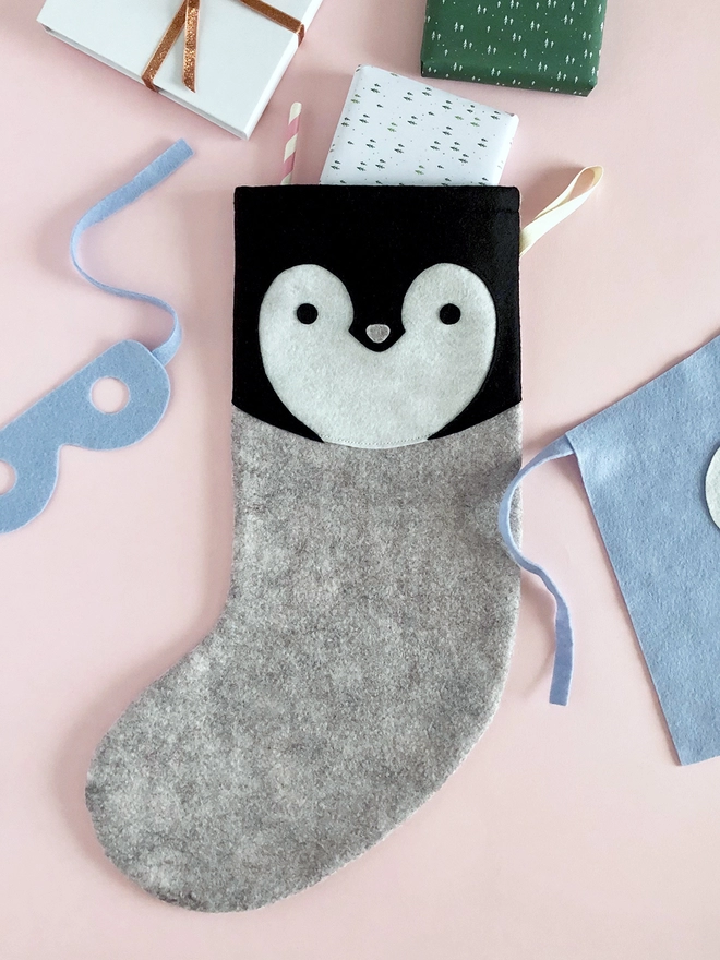 A handmade felt penguin stocking lays on a pink surface with several gifts poking out from the top.