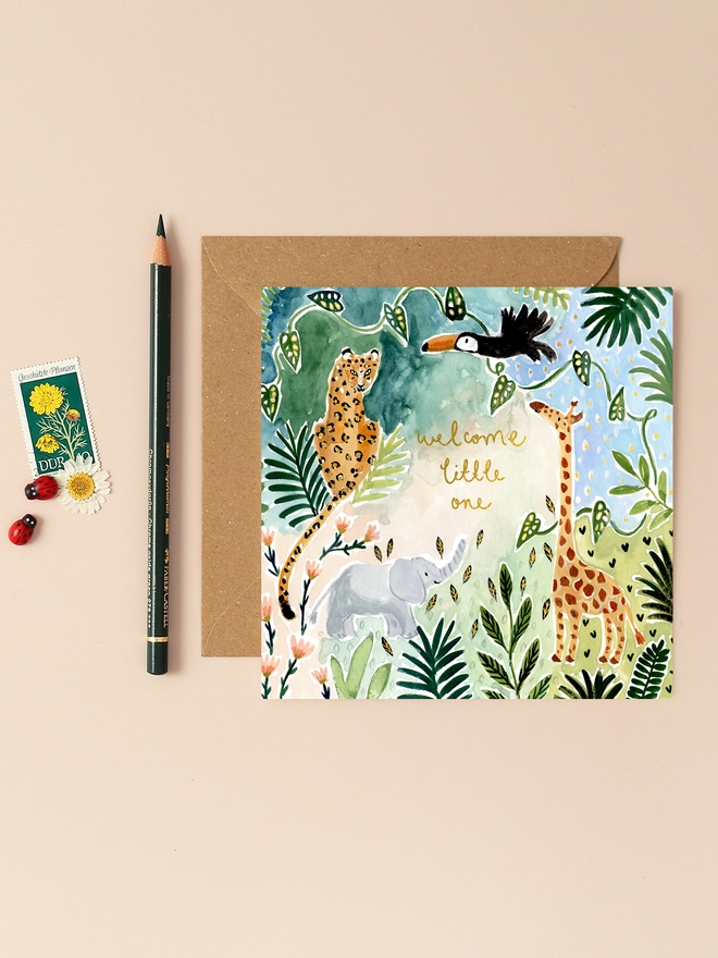 An image of a square greetings card with a colourful jungle themed illustration of a leopard, elephant, toucan and giraffe surrounded by jungle fauna and flora. Elements of the card have been finished with gold foil detailing including a central 'welcome to the world little one' hand written message. The card is seen on top of a brown square kraft envelope.