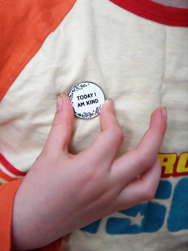A round white enamel pin with a floral design and the words "Today I Am Kind" is pinned to a child's top. Their hand reaches up to hold it.