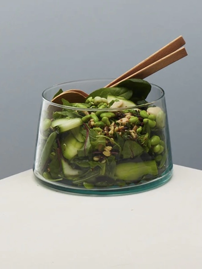 Tall Glass Dish, a tall glass dish sitting on a white surface against a baby blue backdrop. It is filled by a salad. 