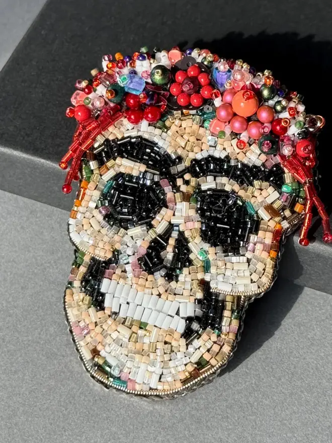 Frida Kahlo Skull Beaded Brooch displayed against a black box, on a grey table. 