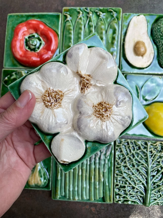Garlic  tile - square, 3D, realistic and glossy. The bright zesty lemon is set off by the lush green glaze of the background. Other fruit and vegetable tiles in the series are on display in the background: fig, savoy cabbage, red capsicum pepper, pear, mange tout, avocado, garlic, asparagus.