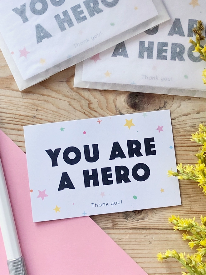 A mini thank you card with the words "You are a hero" and a gentle star design is on a wooden table.