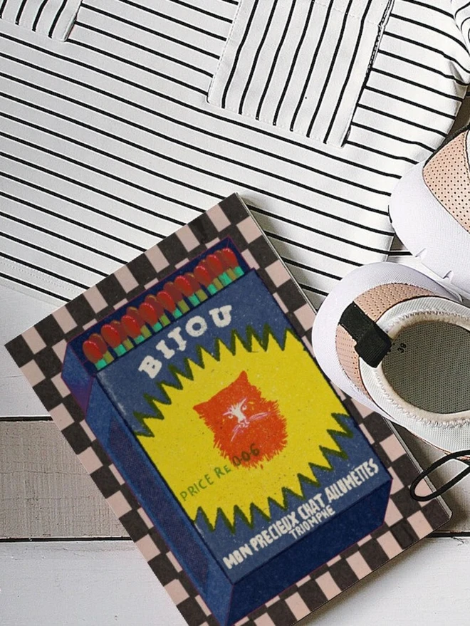 An illustrated vintage-style matchbox labeled “BIJOU.” The box is dark blue with a jagged yellow burst shape in the center, featuring a bright red cat’s face. Text on the burst reads “PRICE Re. 0.06.” Along the bottom, white lettering says “MON PRECIEUX CHAT ALLUMETTES TRIOMPHE.” The top of the matchbox shows red-tipped matches with green stems. The background is a checkered pattern in black and white.