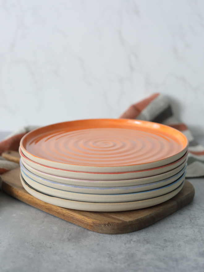 A stack of handmade ceramic lunch plates on a wooden board, topped with a mandarin orange plate with a natural swirl texture, with multicoloured rim edges visible beneath in red, blush, lavender, and blue, styled with a linen cloth against a white marble background