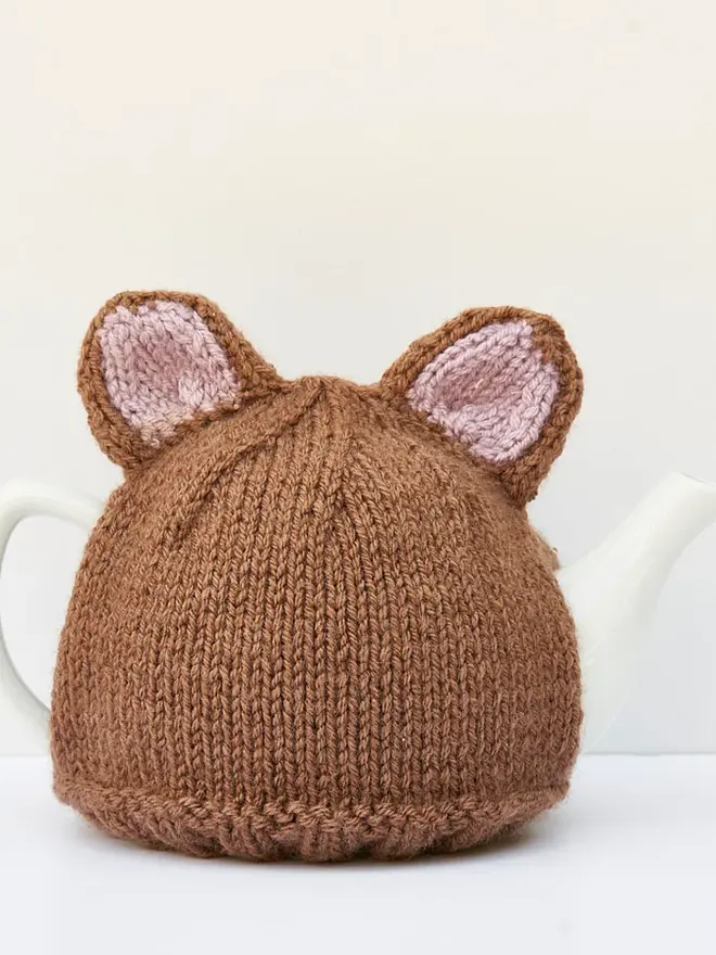 Brown Bear Tea Cosy Teddy Bears Picnic Treat, a brown bear tea pot cover sitting on a white tea pot, on a white surface against a grey backdrop. 