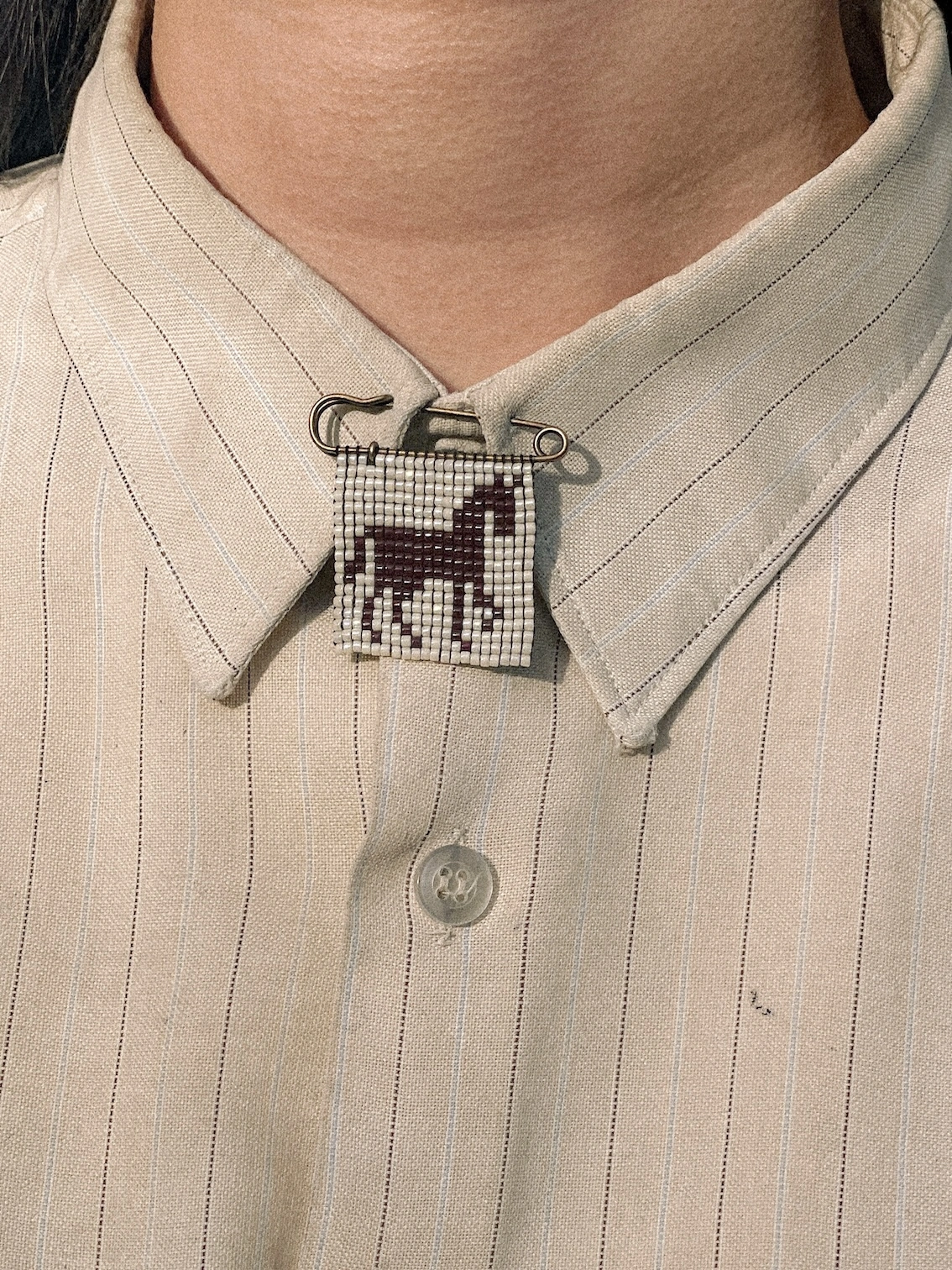 'creature viii' beaded pin. a beaded pin badge with a brown horse silhouette on a pearl background, fastened to a shirt collar.