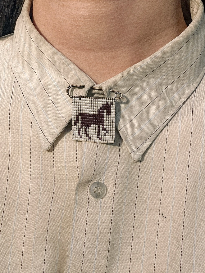 'creature viii' beaded pin. a beaded pin badge with a brown horse silhouette on a pearl background, fastened to a shirt collar.