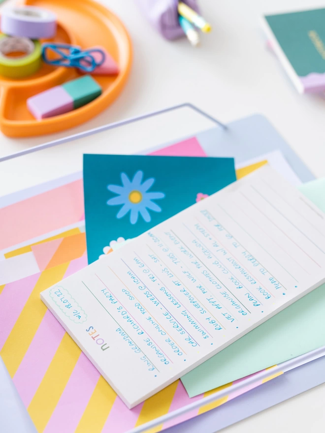 The Raspberry Blossom rainbow coloured lined note pad sits on a desk in a pile of other colourful stationery items from the Happiness collection