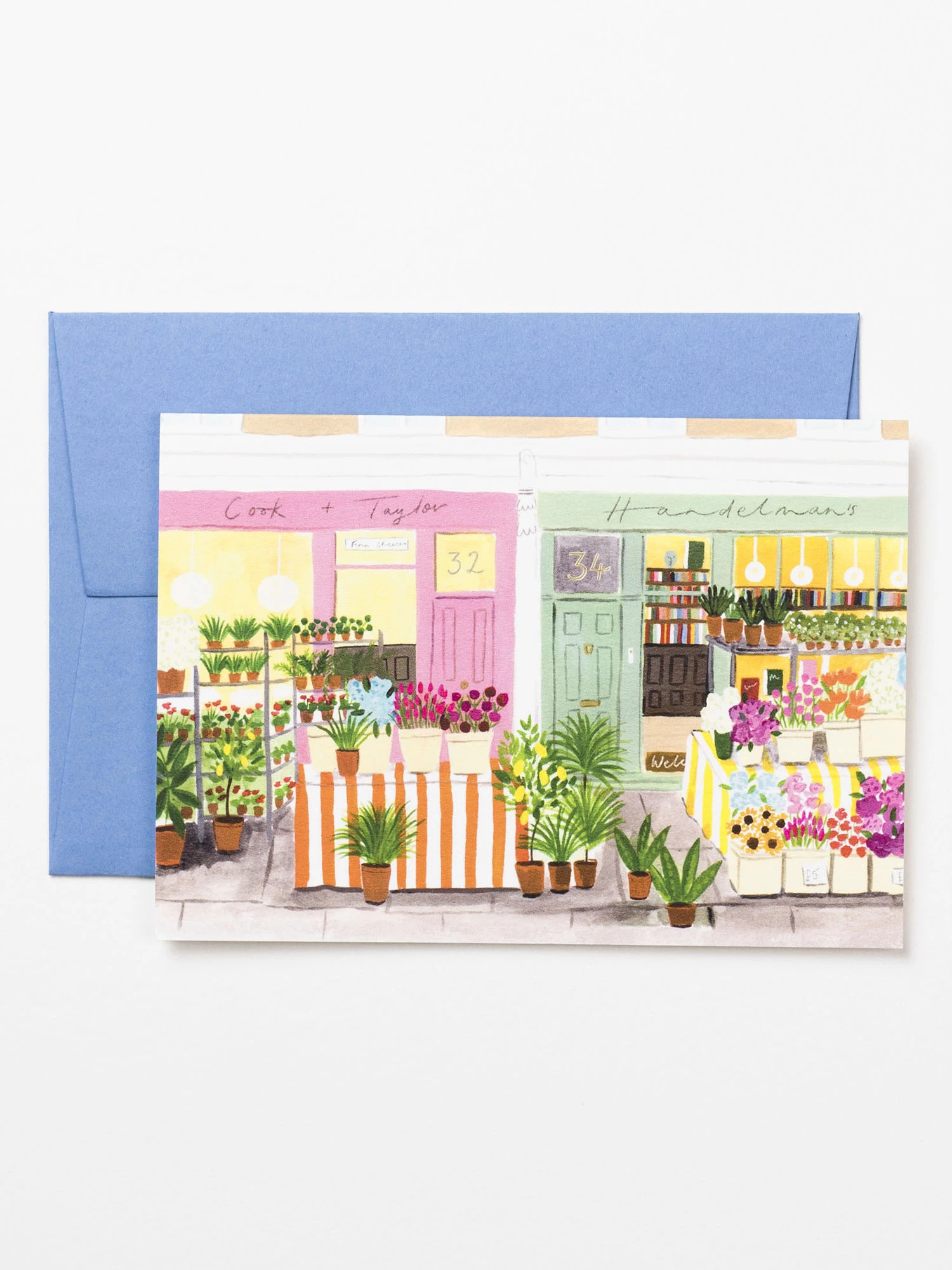 An illustrated card. The card features a painting of a flower market on a street with brightly coloured shop fronts. The flowers are in buckets set on top of tables with bright orange striped tablecloths. The card is placed on top of a pale blue luxury envelope.
