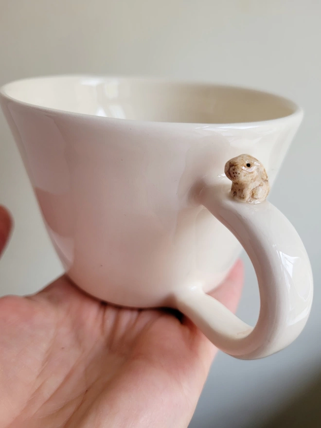 white ceramic cup with a miniature ceramic lop ear bunny rabbit on the handle held in an outstretched hand