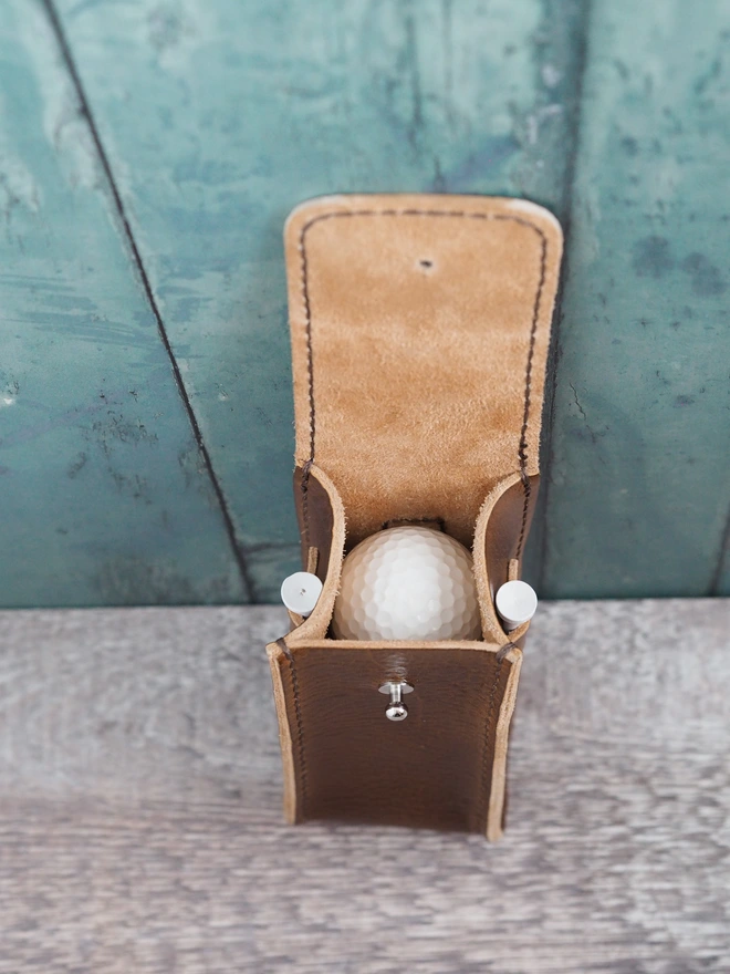 Handmade leather golf ball carry case. The case is open, viewed from above where 3 golf balls are sitting inside stacked. Either side is a single tee in a tee, ready for a game.