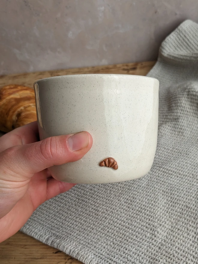 A small white plant pot with croissant design, holding a green leafy plant on a wooden surface.